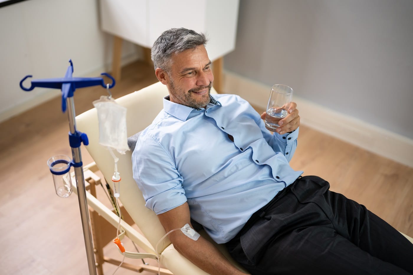 a man in a hospital chair holding a glass of water | Men Holding Glass and Receiving IV Therapy | Synergy Wellness MediSpa in Oceanport, NJ